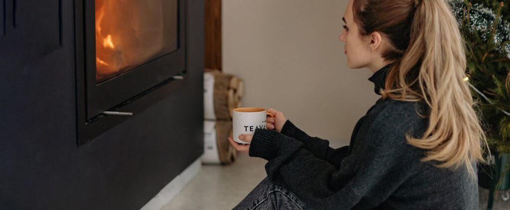 a girl is holding a cup of tea and sit beside the fireplace