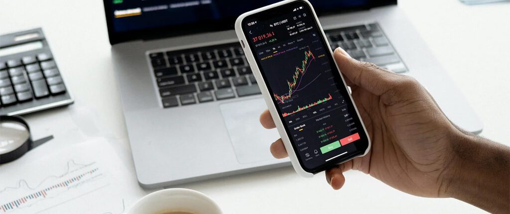 A hand is holding a phone in front of a laptop which showing the chart of stocks.