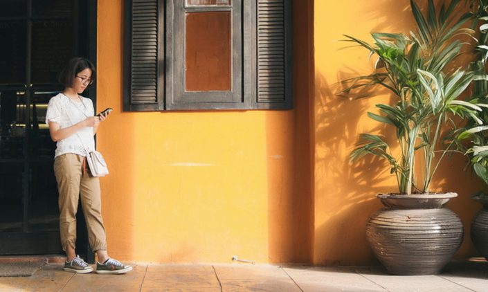 a woman or person waiting in front of a building with yellow wall and plants around her